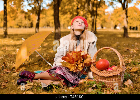 young girl in red cap is sniffing looks romantic up. teenager sitting on the grass holding autumn leaves in his hands. near basket with pumpkin, apple Stock Photo