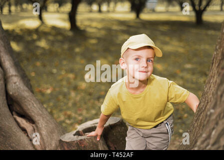 portrait of little boy in yellow cap and t-shirt. kid sits on big tree and smiles Stock Photo
