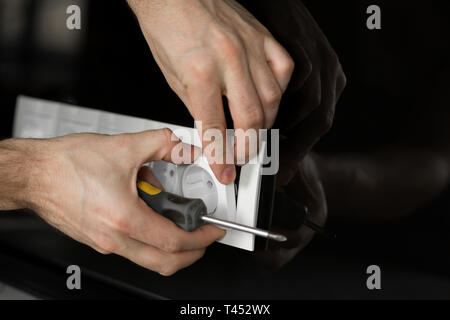 Close-up of an electrician's hand with a screwdriver disassembling a white electrical outlet on a black glass wall. Stock Photo
