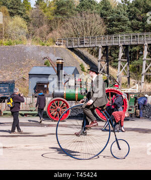 Beamish Museum, Beamish, Durham County, England, United Kingdom, 13 April 2019. Beamish Steam Day: Man in vintage costume riding a penny farthing bicycle with a vintage Clayton & Shuttleworth steam traction engine on display at Beamish Living Museum Stock Photo