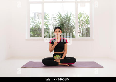 Young asian woman in fitness wear, with vegetarian healthy salad, sitting on floor, against white wall. Stock Photo