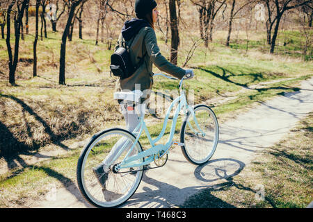 Rear view full height portrait of young woman walking beside bicycle on nature path on sunny spring day. Stock Photo