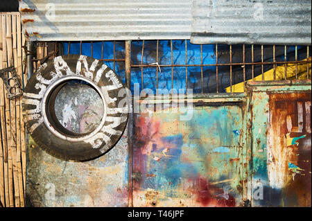 Colorful painted rusty entrance metal gate with an attached old tire as signage. Located behind the gate is an operating vulcanizing business Stock Photo