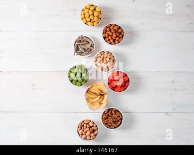 Assortment of different snack for beer, wine, party. Peanuts in coconut glaze, green vasabi, red spicy chilli, yellow cheese glaze, chips, pistachio, crackers, fish on white wooden table. Copy space Stock Photo