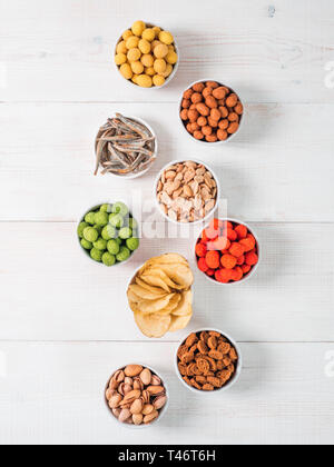 Assortment of different snack for beer,wine,party.Peanuts in coconut glaze,green vasabi,red spicy chilli,yellow cheese glaze and chips,pistachio,crackers,fish on white wooden table.Copyspace.Vertical. Stock Photo
