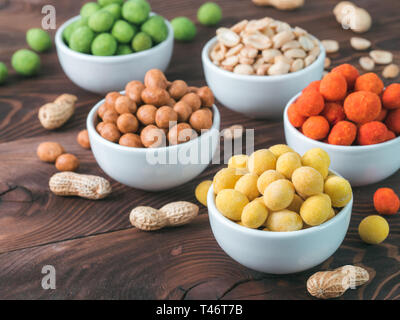 Assortment of different peanuts snack for beer, wine, party bar. Salted peanuts, peanuts in coconut glaze, green vasabi, red spicy chilli, yellow cheese glaze on brown wooden table. Copy space. Stock Photo