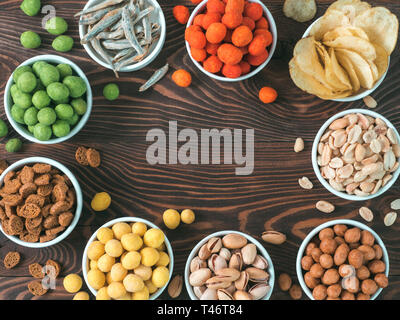 Assortment of different snack for beer, wine, party. Peanuts in coconut glaze, green vasabi, red spicy chilli, yellow cheese glaze, chips, pistachio, crackers, fish on brown wooden table. Copy space Stock Photo