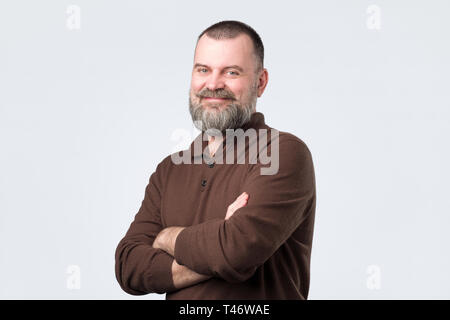 Confident senior man with beard in brown t-shirt crossing hands on chest and looking at camera Stock Photo