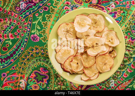 Round green plate with healthy natural homemade apple chips on bright green floral fabric. Fruit healthy snack. Top view, copy space. Stock Photo