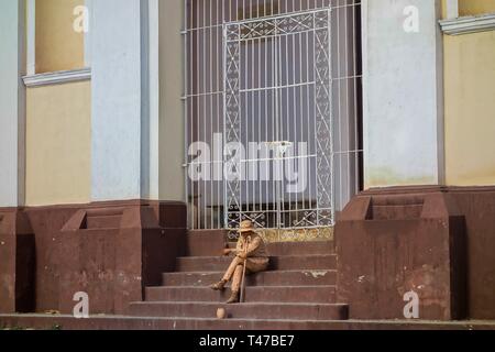 Young Man Busker with Begging Bowl covered in Paint sitting on stairs of Iglesia y Convento de San Francisco Church in Trinadad, Cuba Stock Photo