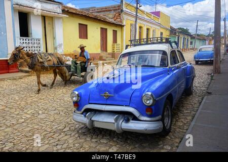 Old Man in Horse Carriage and Classic Cuban Taxi Car on Cobblestone Street showing contrasting Transport Modes and typical City Life in Trinidad, Cuba Stock Photo