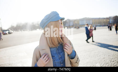 Stylish young blonde in denim cap and beige raincoat. Beautiful young woman walking on the street, Sunny day. Stock Photo