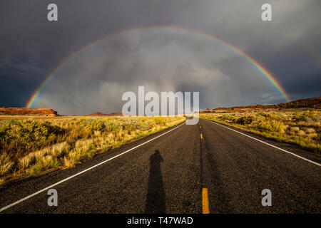 The shadow of the photographer shows on the road below a full rainbow and thuderstorm clouds over Scenic Highway 211 in Canyonlands National Park, Sou Stock Photo