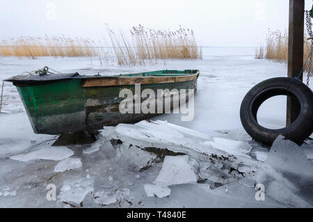 Winter landscape with old wooden boat on crushed ice, of frozen lake next to bridge. Withered reed and horizon in background. Stock Photo