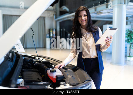 Beautiful woman in dealership center staying near new car handing tablet. Shows functionality of the car Stock Photo