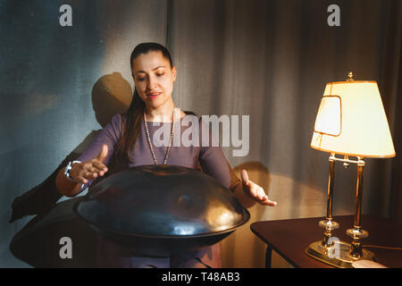 Young woman try playing on a first generationan instrument called '' Hang '' or ''Hang drum'' in the room with retro lamp Stock Photo