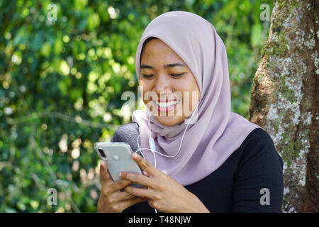 Close-up shot of muslim lady in hijab using smartphone with earphone. Footage of Malaysian young woman or student looking at smartphone. Stock Photo