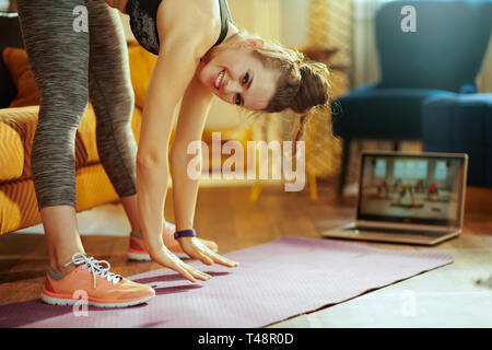 smiling young sports woman in sport clothes at modern home using laptop to watch fitness streaming on internet while doing stretching on fitness mat. Stock Photo