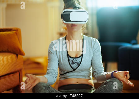 Portrait of relaxed young sports woman in fitness clothes in the modern house doing yoga in VR gear. Stock Photo