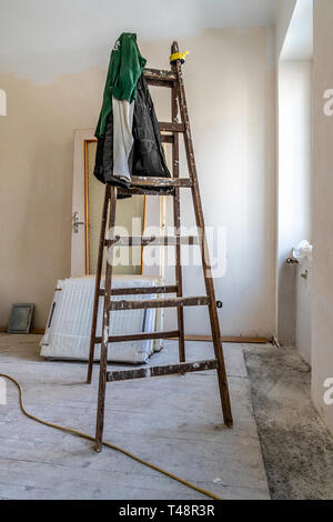 Empty Wooden Ladder with Hanging Dirty Green Colored Pullover Clothes in a White Color Painted Wall Apartment Room Under Construction next to the Wind Stock Photo