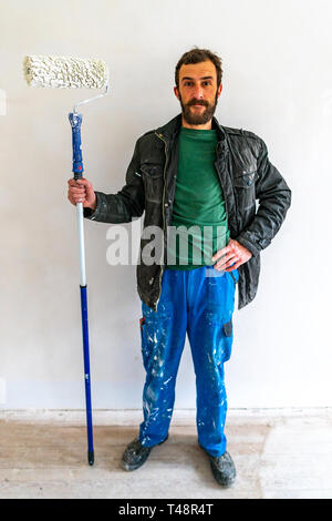 Young Caucasian Brown Haired Man with Beard and Blue Colored Dungarees Working Trousers Standing and Holding his Fabric Paint Stick Roller Extension P Stock Photo