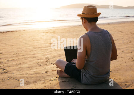 Back view of barefoot man in hat sitting on sandy beach near waving sea and browsing modern laptop Stock Photo