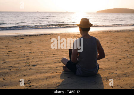 Back view of barefoot man in hat sitting on sandy beach near waving sea and browsing modern laptop Stock Photo