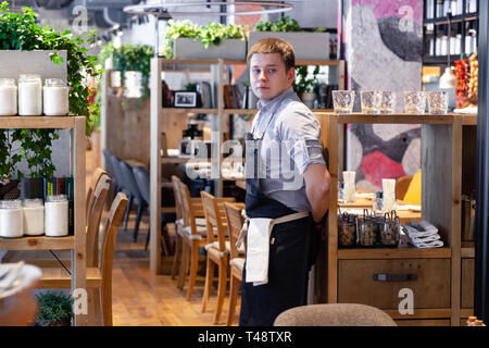 Russia Novosibirsk 2018-11-26 a young male waiter in uniform, shirt and black apron is standing in the restaurant and watching the sales area and cust Stock Photo