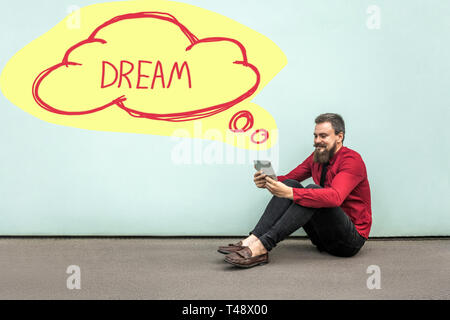 Happy bearded businessman in red shirt sitting on floor, holding, looking at his digital tablet and dreaming, yellow cloud shape on wall with dream te Stock Photo