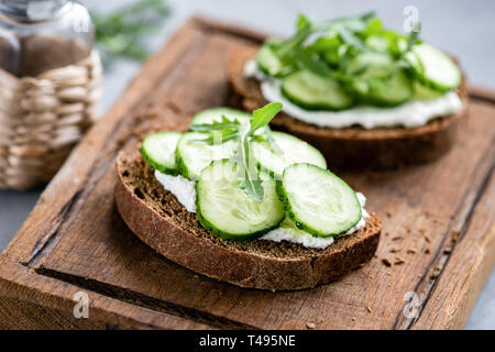 Rye bread with cream cheese and cucumber on wooden cutting board. Closeup view, selective focus, horizontal orientation Stock Photo