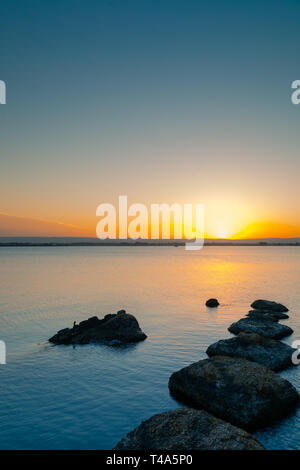Sunset over the calm water, rocks in the foreground Stock Photo