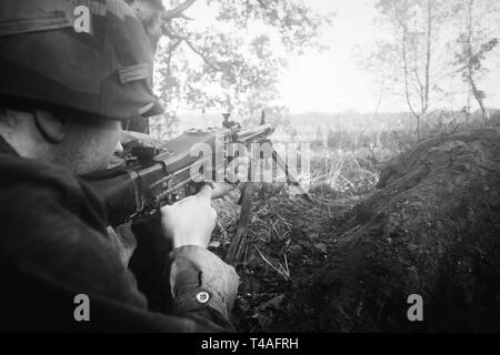Hidden Re-enactor Dressed As German Wehrmacht Infantry Soldier In World War II Aiming a Machine Gun From Trench In Misty Forest. WWII WW2 Weapon MG 42 Stock Photo