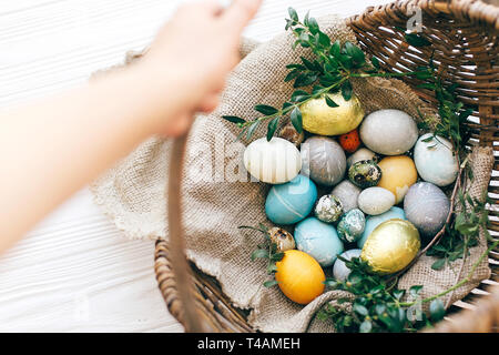 Hand holding  rustic wicker basket with stylish Easter eggs with green buxus branches on white wooden background. Easter hunt concept. Happy Easter. Stock Photo