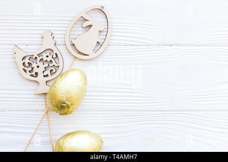 Stylish Easter chocolate eggs in golden foil and simple wooden bunny and chicken decorations on white wooden background, flat lay with space for text. Stock Photo