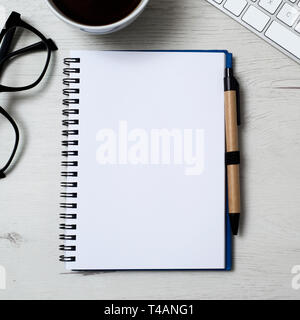 high angle view of a notebook with a blank space, a pen, a pair of black plastic rimmed eyeglasses, a cup of coffee and a keyboard, on a white wooden  Stock Photo