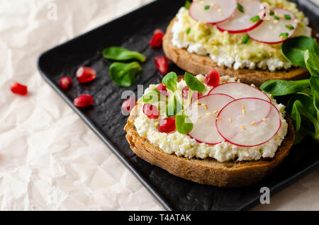 Toasts with cottage cheese and smashed avocado, radish, corn salad plant and pomegranate seeds on black plate. Top view. Copy space. Healthy eating an Stock Photo
