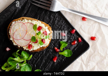Toast with cottage cheese and smashed avocado, radish, corn salad plant and pomegranate seeds on black plate. Top view. Copy space. Healthy eating and Stock Photo