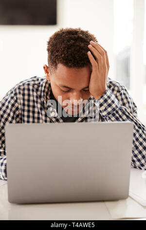 Depressed black teenage boy with head in hands using a laptop computer at home, vertical