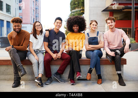 Six young adult friends sitting in a row on a bench in a city street smiling to camera, full length Stock Photo