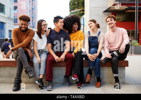 Six young adult friends sitting in a row on a bench in the street looking at each other, full length Stock Photo