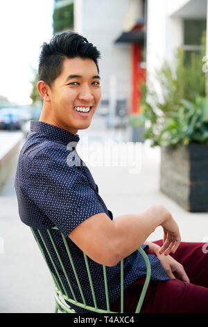 Millennial Asian man sitting on a chair in a city street, turning and smiling to camera, close up Stock Photo