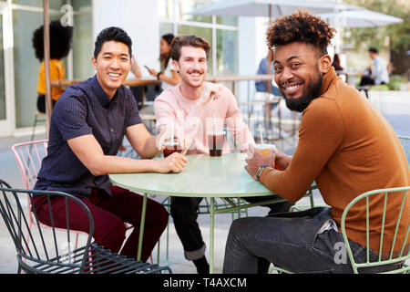 Three millennial male friends talking over cold brews outside a cafe in a city street turn to camera Stock Photo