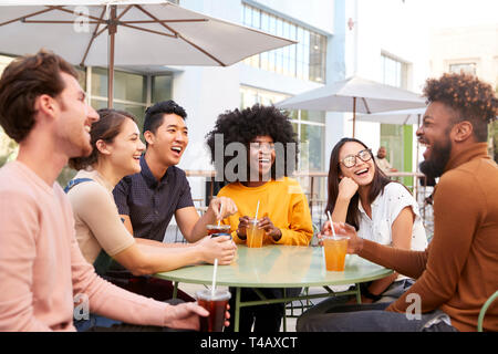 Six trendy millennial friends sit drinking cold brews talking and laughing outside a cafe Stock Photo