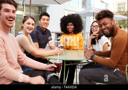 Six trendy millennial friends sitting drinking outside a cafe, turning and smiling to camera Stock Photo
