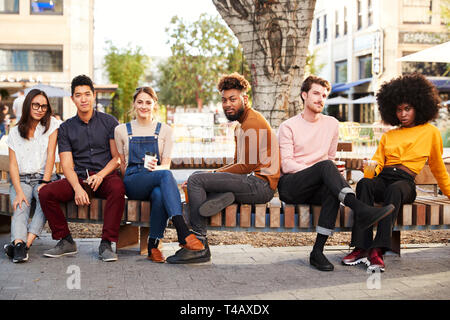 Six millennial hipster friends in the city relaxing by a fountain with drinks, smiling to camera Stock Photo