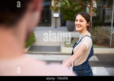 Over shoulder view of millennial couple crossing the road in the city holding hands, selective focus Stock Photo