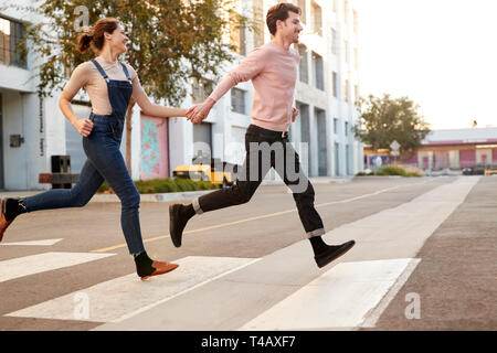 Millennial hipster couple running across the road in the city holding hands, side view Stock Photo