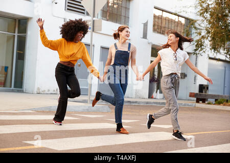 Three millennial hip girlfriends holding hands and laughing as they run across a pedestrian crossing Stock Photo