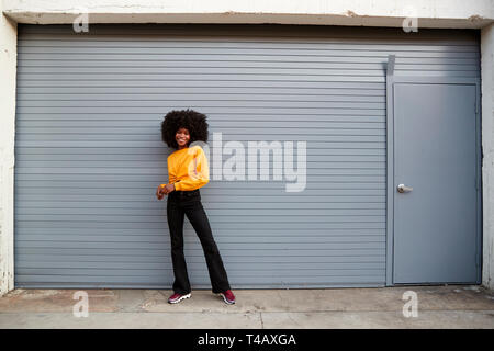 Hip young black woman standing against grey security shutters smiling to camera, full length Stock Photo