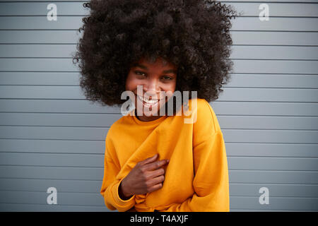 Young black woman with afro against grey wall, raising shoulder, smiling to camera, close up Stock Photo